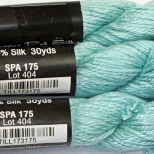 May include: Close-up of three skeins of light teal-colored silk thread. Each skein has a black label with white text that reads "100% Silk 30 yds", "SPA 175 Lot 404", and a barcode. The thread is soft and lustrous.
