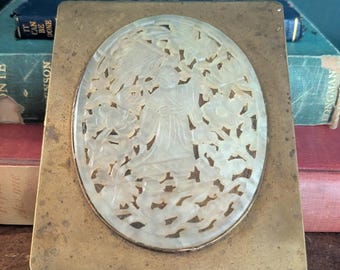 Puede incluir: Caja de latón antigua con incrustación ovalada de nácar tallado. La incrustación presenta un relieve detallado de una figura entre elementos florales. La caja es rectangular con tapa abisagrada, mostrando una estética vintage.