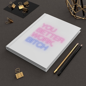 May include: A white journal with the text "YOU BETTER WORK, BITCH" in pink and blue neon-style lettering. Gold and black pencils and gold binder clips are scattered around the journal on a dark gray surface.