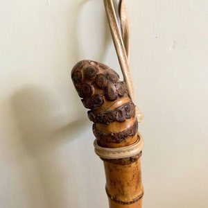 May include: A carved wooden walking stick with a leather strap. The stick is brown and has a detailed design. The strap is beige.