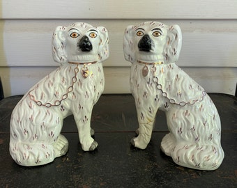 May include: Two white porcelain dog figurines with brown spots and gold chains around their necks. The dogs are sitting with their heads turned to the side.