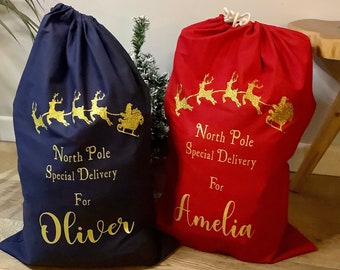 May include: Two Christmas gift bags, one red and one blue, with gold glitter reindeer and Santa Claus designs. The bags are personalized with the names "Amelia" and "Oliver" and the text "North Pole Special Delivery".