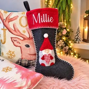 May include: A dark grey felt Christmas stocking with a red cuff featuring the white text Millie. The stocking is decorated with a gnome applique wearing a red hat and holding a red and white chequered heart. It has a black hanging loop.