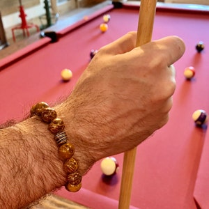 May include: A brown wooden beaded bracelet with silver accents. The bracelet is worn on a person's wrist, and the person is holding a pool cue.