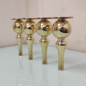 May include: Four gold-colored furniture legs. Each leg has a rounded, bulbous top with a decorative, flat piece connecting them. The legs taper down to a slender, fluted base. The polished surface reflects the surrounding environment.