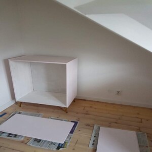 May include: A white cabinet with gold legs, sitting on a wooden floor. Two white pieces of wood are on the floor in front of the cabinet.