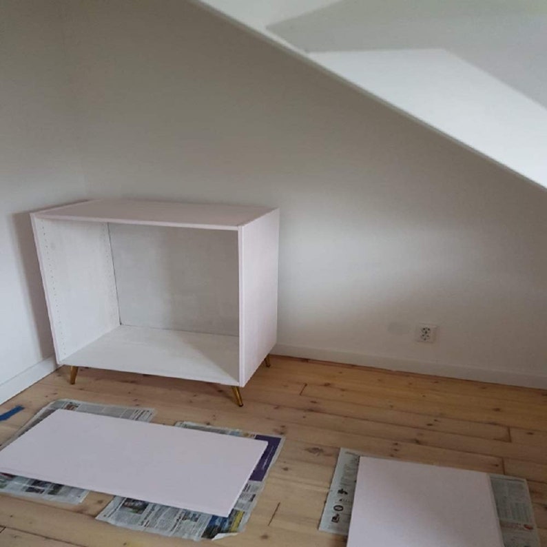 May include: A white cabinet with gold legs, sitting on a wooden floor. Two white pieces of wood are on the floor in front of the cabinet.