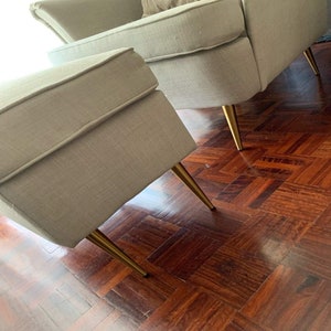 May include: A light gray upholstered ottoman with gold metal legs. The ottoman is sitting on a dark wood floor.