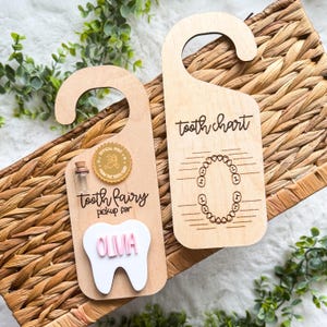 May include: Two wooden tooth fairy door hangers. One has a tooth-shaped cutout with the name "OLIMA" in pink, a small bottle, and text that reads "tooth fairy pickup for." The other says "tooth chart."