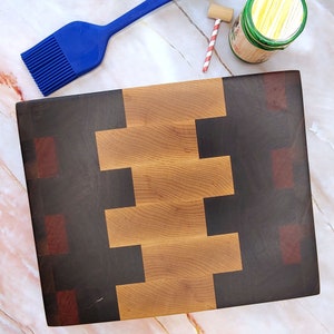 May include: A wooden cutting board with a checkerboard pattern of light and dark wood. The board has a light brown center with dark brown and reddish brown squares around the edges.