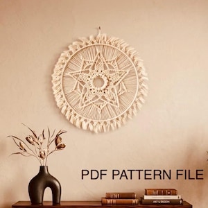 May include: A cream-colored macrame wall hanging with a starburst design is displayed on a beige wall. Below, a dark wooden console table with gold legs holds a black vase with dried stems and books. The text "PDF PATTERN FILE" is visible.