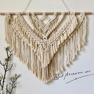 May include: A large, off-white macrame wall hanging with a wooden dowel at the top. The macrame is in a triangular shape with a fringe at the bottom. The macrame is made with a thick, natural fibre cord. The wall hanging is signed "Just Macrame.com" in black ink.