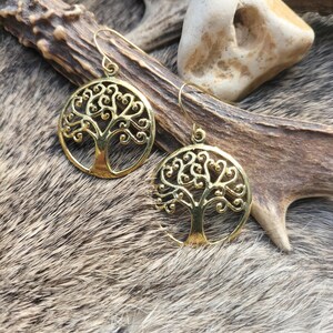 May include: Two gold-colored earrings with a tree of life design. The earrings are circular with intricate, swirling branches and a trunk. The earrings hang from a simple hook. The earrings are displayed on a furry, brown and gray surface.