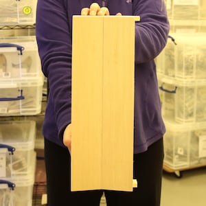 Puede incluir: Un panel de madera de color claro, sostenido verticalmente. El panel está hecho de dos piezas de madera unidas, con un pequeño bloque de madera adjunto en la parte inferior. La madera tiene una veta natural y un acabado liso. El panel es sostenido por una persona.