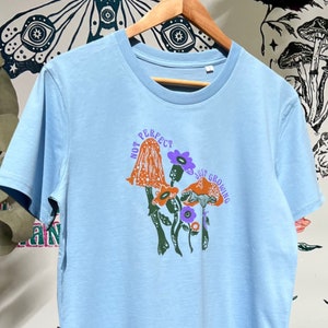May include: Light blue t-shirt featuring a graphic of mushrooms and flowers. The text "Not Perfect, Just Growing" is printed on the shirt.