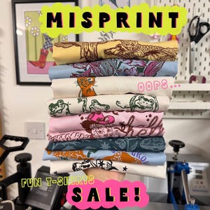 以下が含まれることがあります： 猫、カエル、カウボーイハットなど、さまざまなグラフィックデザインが施された折り畳まれたTシャツの山。 「MISPRINT」と「FUN T-SHIRTS SALE！」の文字が鮮やかな色で表示されています。 シャツは、黄色、青、ピンクなど、さまざまな色があります。