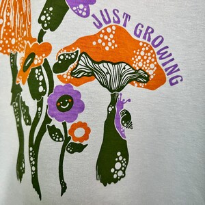 May include: White t-shirt with a green, orange and purple mushroom graphic and the text "Just Growing".