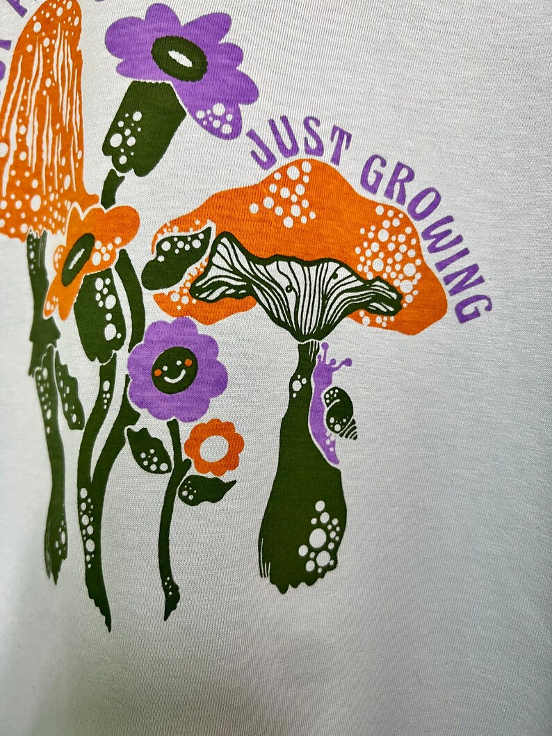 May include: White t-shirt with a green, orange and purple mushroom graphic and the text "Just Growing".