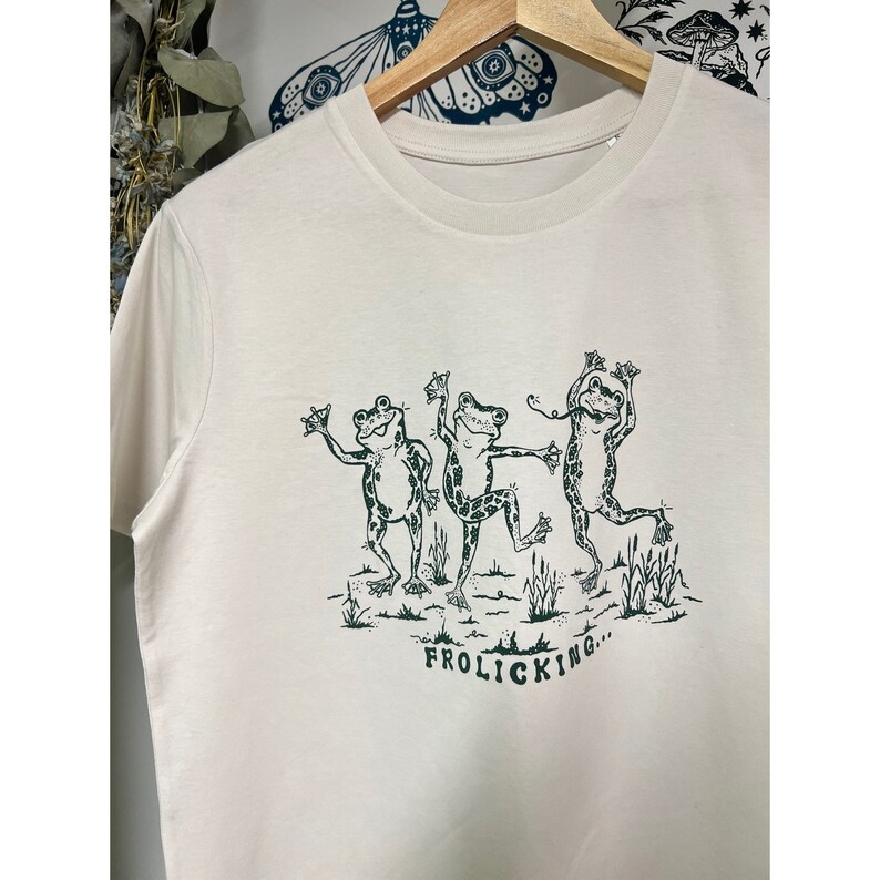 May include: A cream-colored t-shirt with a green illustration of three frogs dancing with the text "FROLICKING..." underneath.