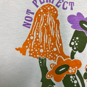 May include: A white t-shirt featuring a graphic print of orange and green mushrooms and purple flowers. The text "NOT PERFECT" is printed in purple above the mushrooms.