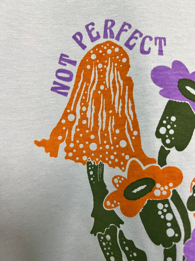 May include: A white t-shirt featuring a graphic print of orange and green mushrooms and purple flowers. The text "NOT PERFECT" is printed in purple above the mushrooms.