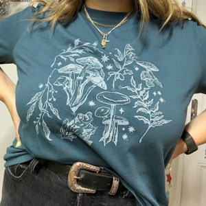 May include: A teal blue t-shirt with a white graphic print of mushrooms, a frog, and stars. The graphic is centred on the shirt.