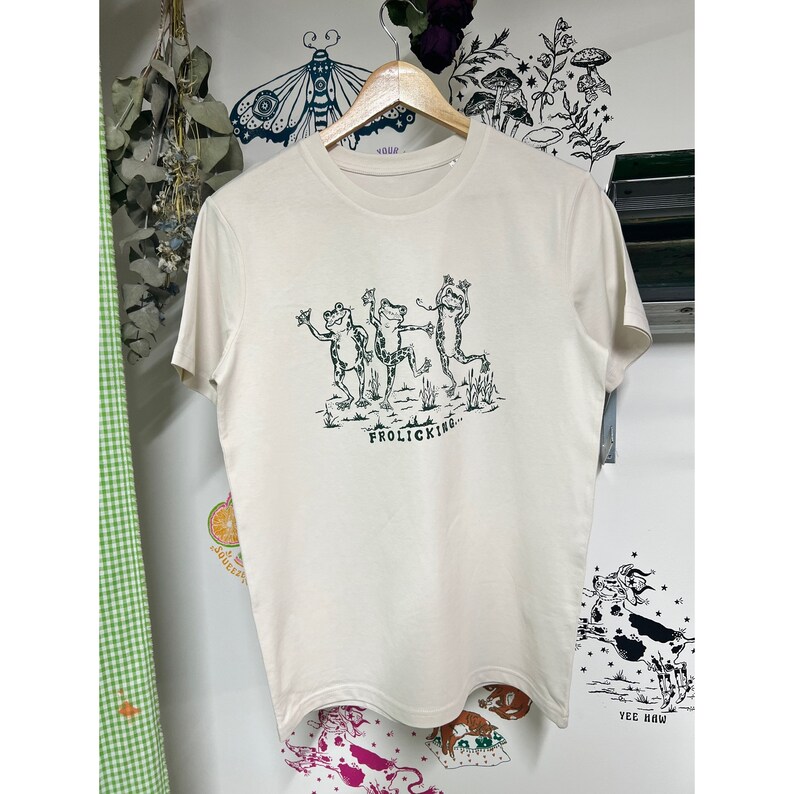 May include: A cream-colored t-shirt with a green graphic of three frogs dancing and the text "Frolicking..." printed on the front.