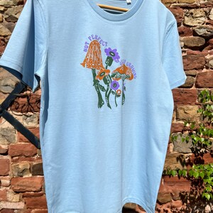 May include: A light blue t-shirt featuring a graphic of mushrooms and flowers. The text "Not Perfect, Just Growing" is printed on the shirt.
