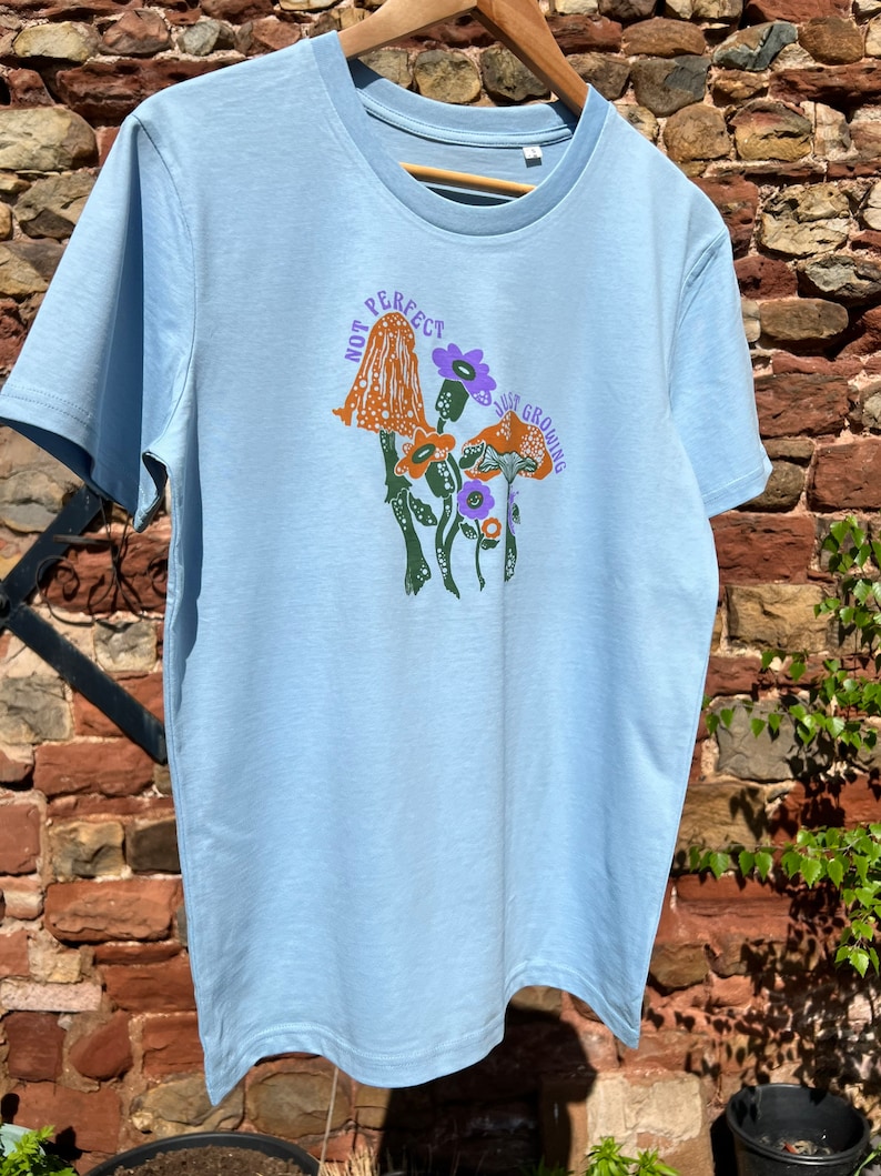 May include: A light blue t-shirt featuring a graphic of mushrooms and flowers. The text "Not Perfect, Just Growing" is printed on the shirt.