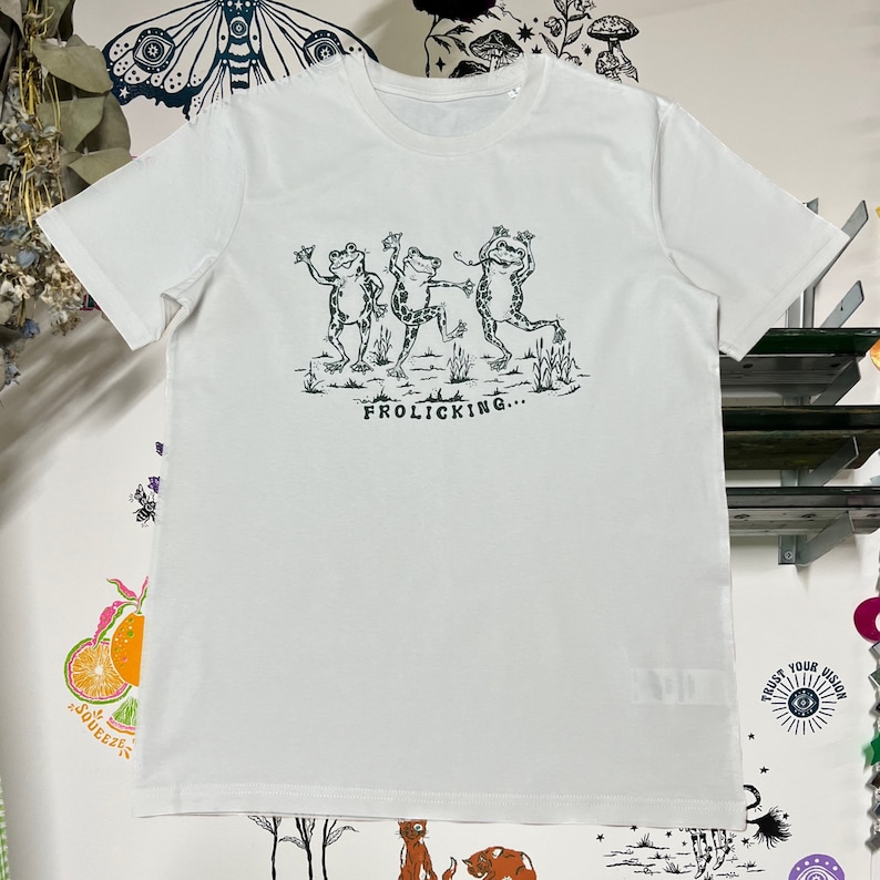 May include: White t-shirt with a graphic of three frogs dancing and the text "Frolicking..." printed in black.