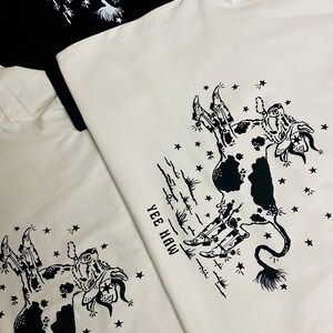 May include: Two white t-shirts with a black and white illustration of a cow wearing cowboy boots and a star-filled sky. The text "Yee Haw" is printed on the shirts.