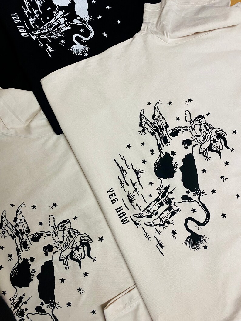 May include: Two white t-shirts with a black and white illustration of a cow wearing cowboy boots and a star-filled sky. The text "Yee Haw" is printed on the shirts.