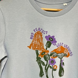 May include: Light blue t-shirt featuring a graphic of mushrooms and flowers. The text "Not Perfect Just Growing" is printed on the shirt.