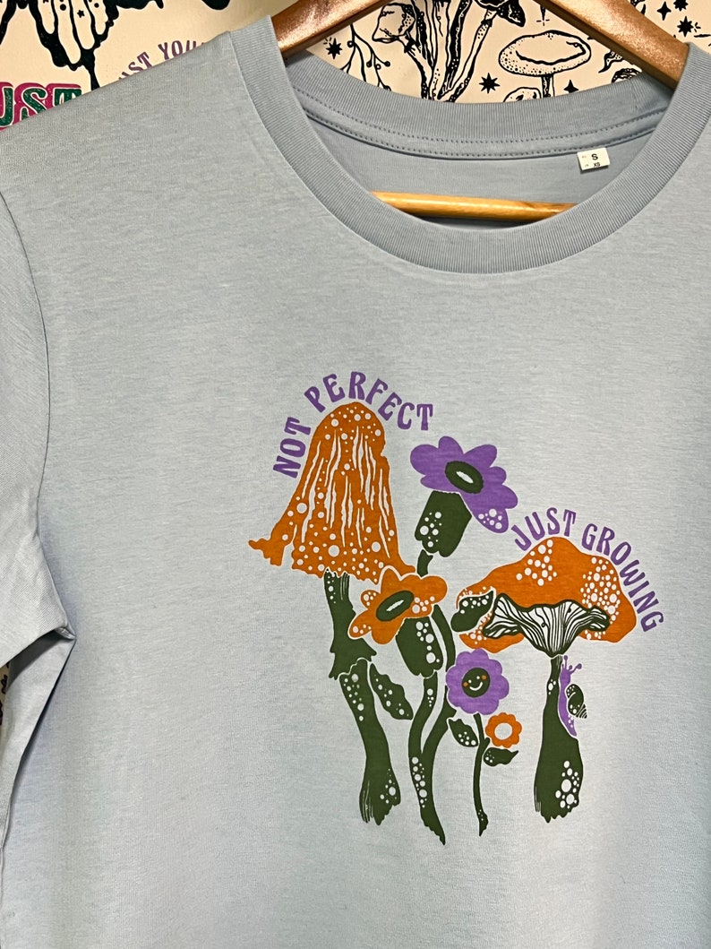 May include: Light blue t-shirt featuring a graphic of mushrooms and flowers. The text "Not Perfect Just Growing" is printed on the shirt.