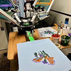 May include: A light blue t-shirt with a screen printed design of mushrooms and flowers. The text "PERFECT" and "JUST GROWING" is printed on the shirt. The shirt is being printed on a screen printing press.