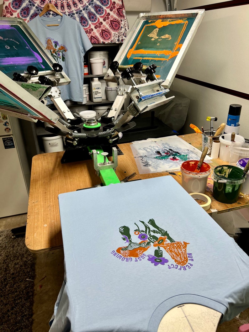 May include: A light blue t-shirt with a screen printed design of mushrooms and flowers. The text "PERFECT" and "JUST GROWING" is printed on the shirt. The shirt is being printed on a screen printing press.