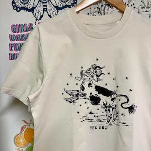 May include: A cream colored t-shirt with a black and white illustration of a cow wearing a cowboy hat and boots. The cow is surrounded by stars and the text "Yee Haw" is printed below the cow.