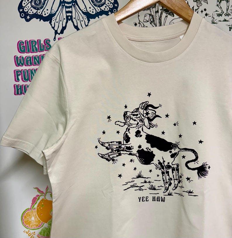 May include: A cream colored t-shirt with a black and white illustration of a cow wearing a cowboy hat and boots. The cow is surrounded by stars and the text "Yee Haw" is printed below the cow.