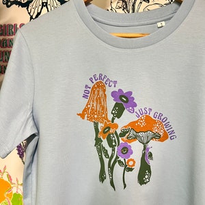 May include: Light blue t-shirt featuring a graphic of mushrooms and flowers. The text "Not Perfect Just Growing" is printed on the shirt.