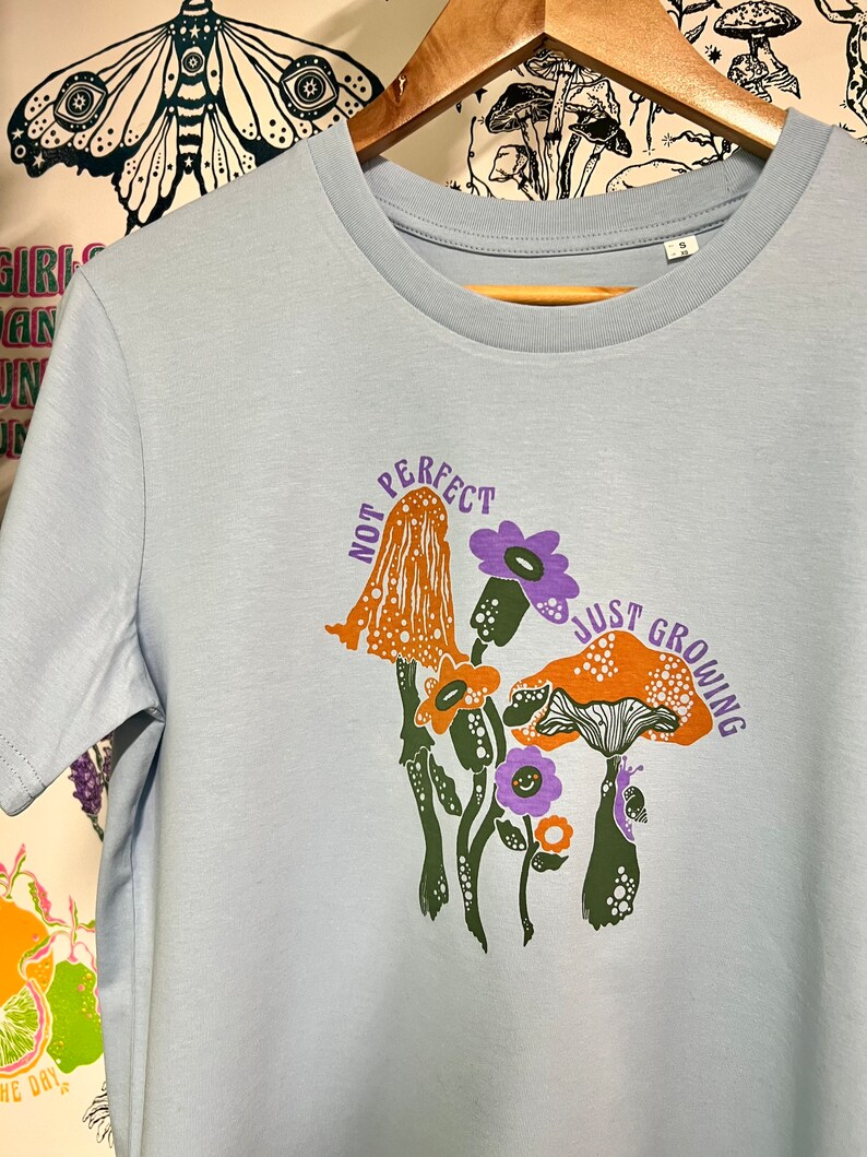 May include: Light blue t-shirt featuring a graphic of mushrooms and flowers. The text "Not Perfect Just Growing" is printed on the shirt.
