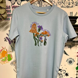 May include: Light blue t-shirt featuring a graphic of mushrooms and the text "Not Perfect Just Growing".