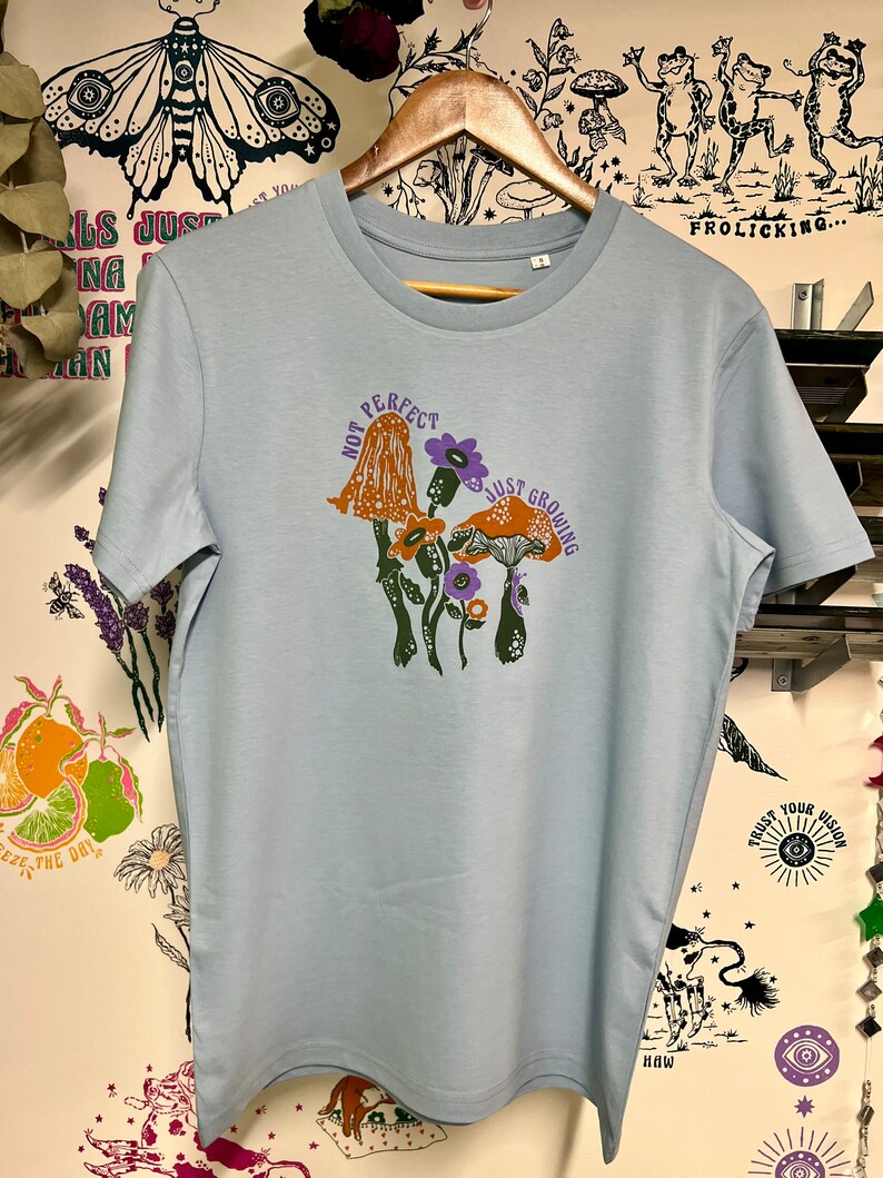 May include: Light blue t-shirt featuring a graphic of mushrooms and the text "Not Perfect Just Growing".