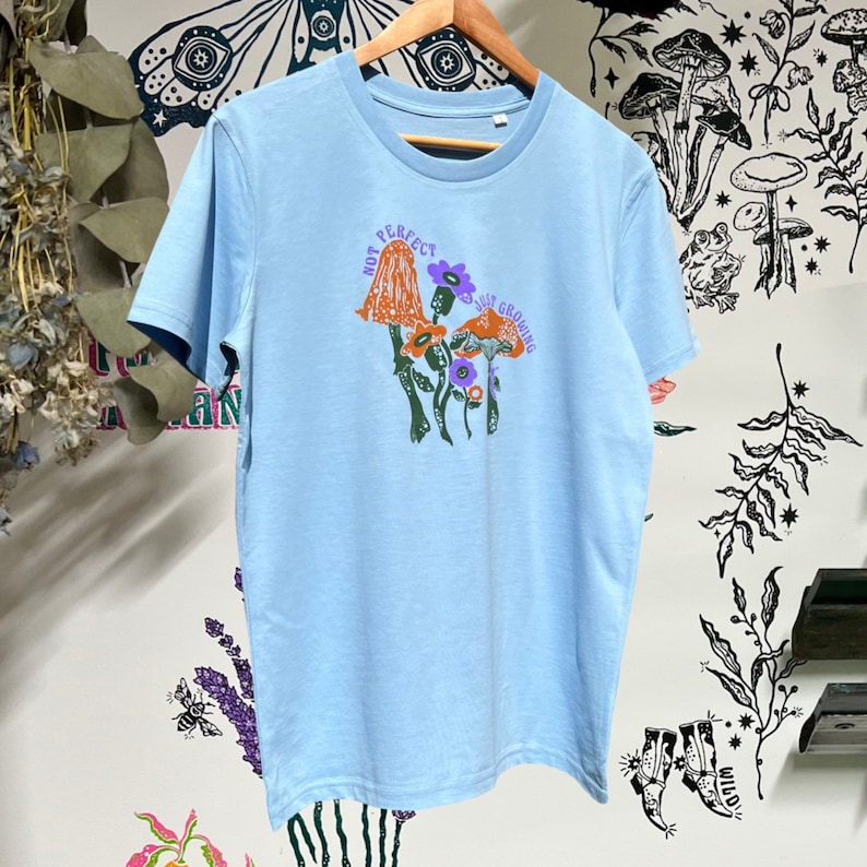 May include: Light blue t-shirt featuring a graphic of mushrooms and flowers. The text "Not Perfect, Just Growing" is printed on the shirt.