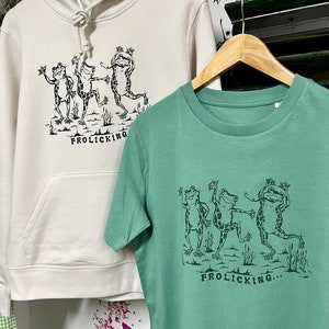 May include: Two shirts with a graphic of three frogs dancing and the text "Frolicking..." printed on them. The shirt on the left is a cream-colored hoodie and the shirt on the right is a green t-shirt.