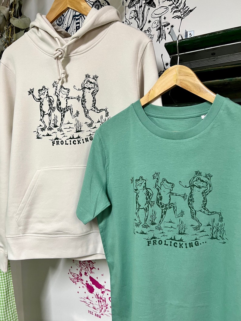 May include: Two shirts with a graphic of three frogs dancing and the text "Frolicking..." printed on them. The shirt on the left is a cream-colored hoodie and the shirt on the right is a green t-shirt.