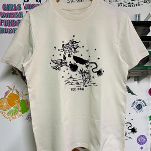 May include: A cream colored t-shirt with a black and white illustration of a cow jumping with stars around it. The text "Yee Haw" is printed below the cow.
