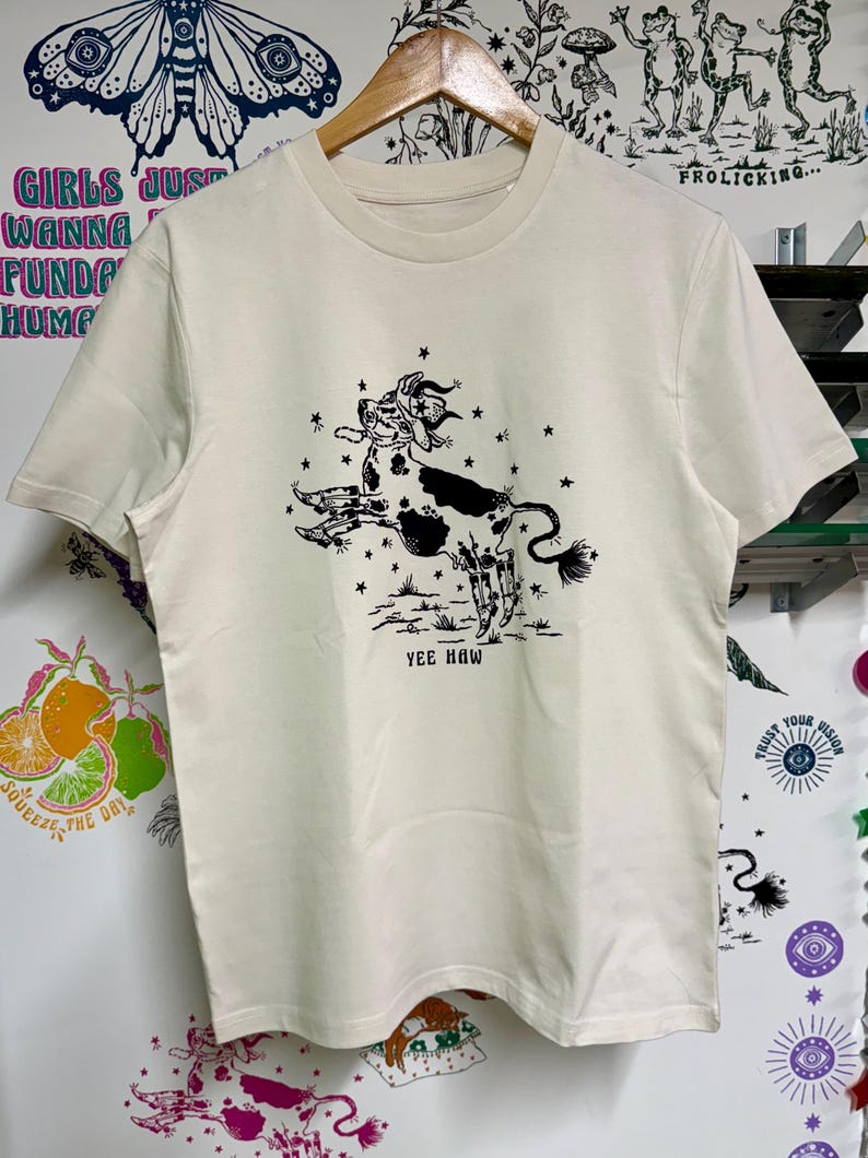 May include: A cream colored t-shirt with a black and white illustration of a cow jumping with stars around it. The text "Yee Haw" is printed below the cow.