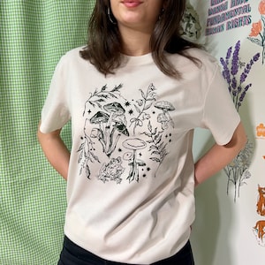 May include: A cream-coloured t-shirt with a black and green print of mushrooms, a frog, and stars.