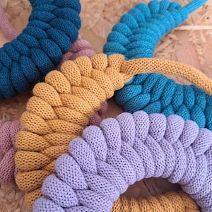 Puede incluir: Primer plano de varios artículos trenzados y circulares de punto en varios colores. Los colores incluyen turquesa, azul claro, amarillo mostaza, lavanda y un rosa apagado. Los artículos parecen estar hechos de un hilo suave y texturizado.