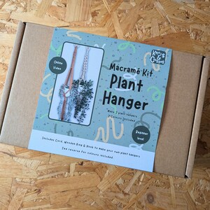 May include: A rectangular brown cardboard box featuring a blue label for a Macramé Kit Plant Hanger. The label displays two finished plant hangers, the text Knots & Crosses, and notes that it is a beginner level kit to make two hangers.